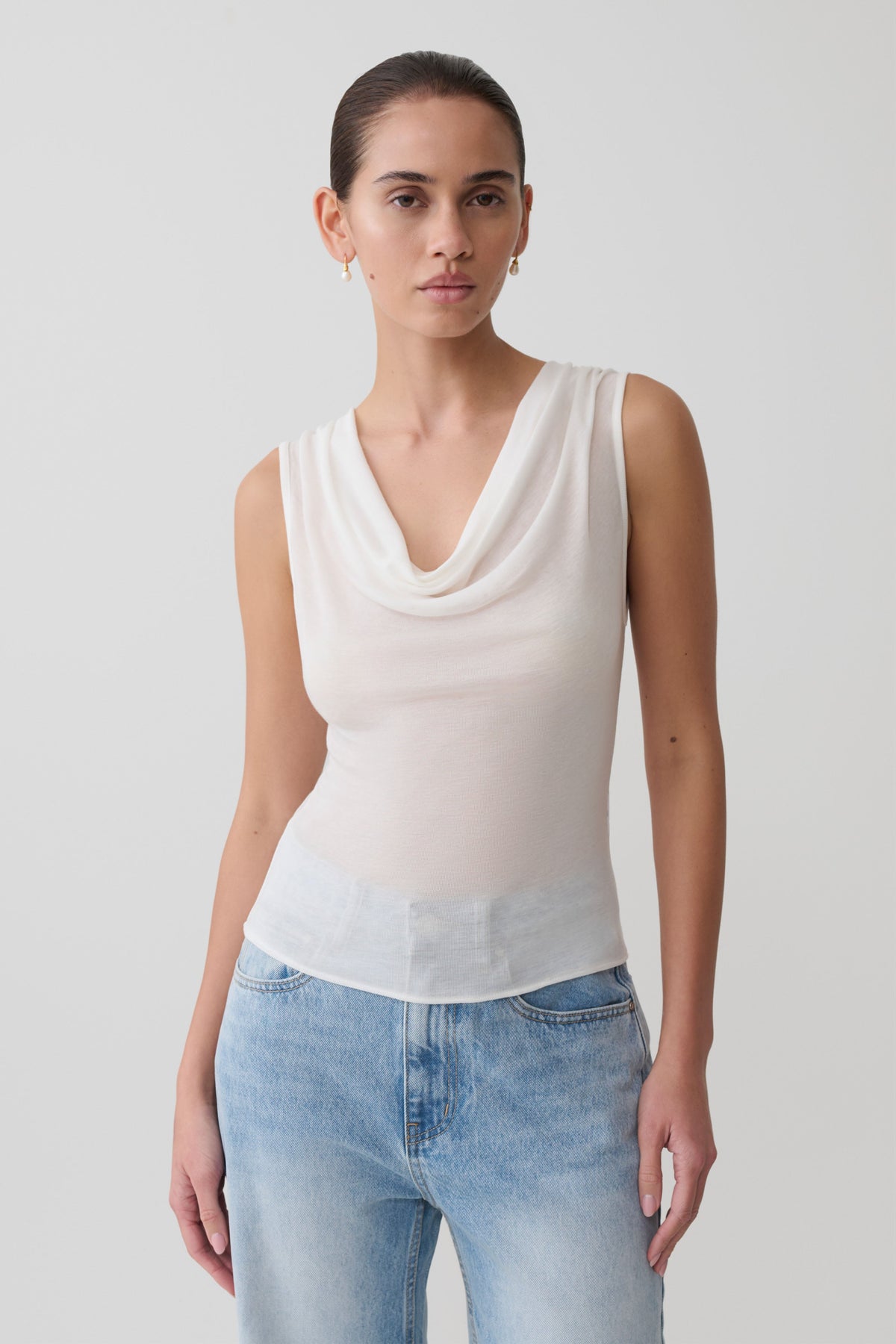 Woman wearing a white sleeveless sheer top and denim pants.