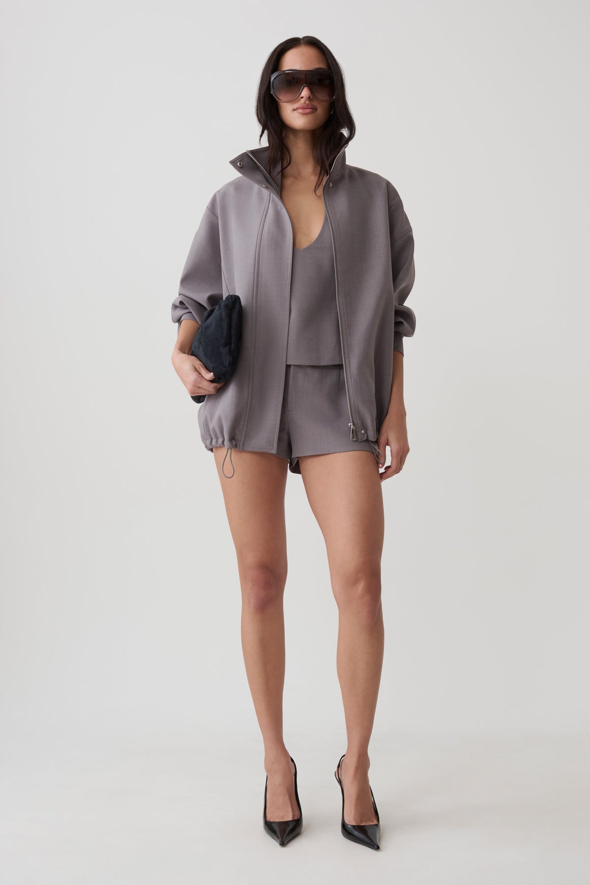 A model stands against a light gray studio background wearing a coordinated gray outfit consisting of an oversized zip-front jacket, matching top, and shorts. She is also wearing black pointed heels and holding a small dark clutch.