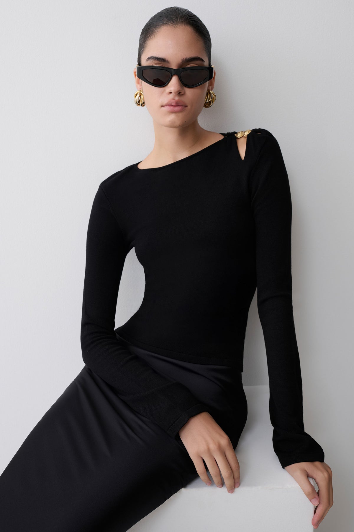 Woman wearing a black long-sleeve dress and sunglasses against a white background.