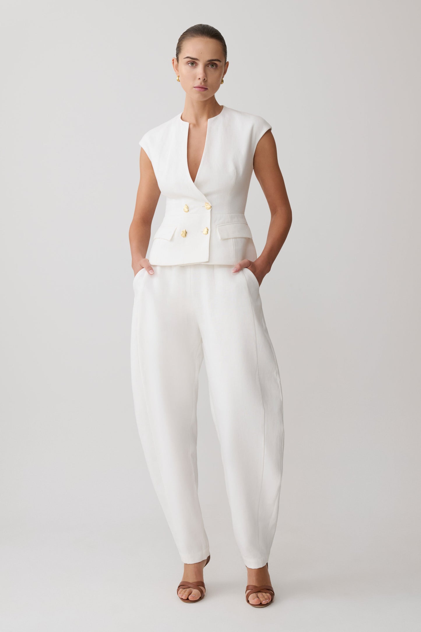 Woman wearing a white sleeveless linen vest with gold buttons with matching pants.