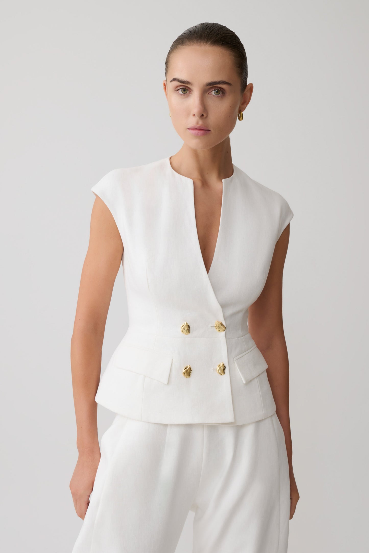 Woman wearing a white sleeveless linen vest with gold buttons.