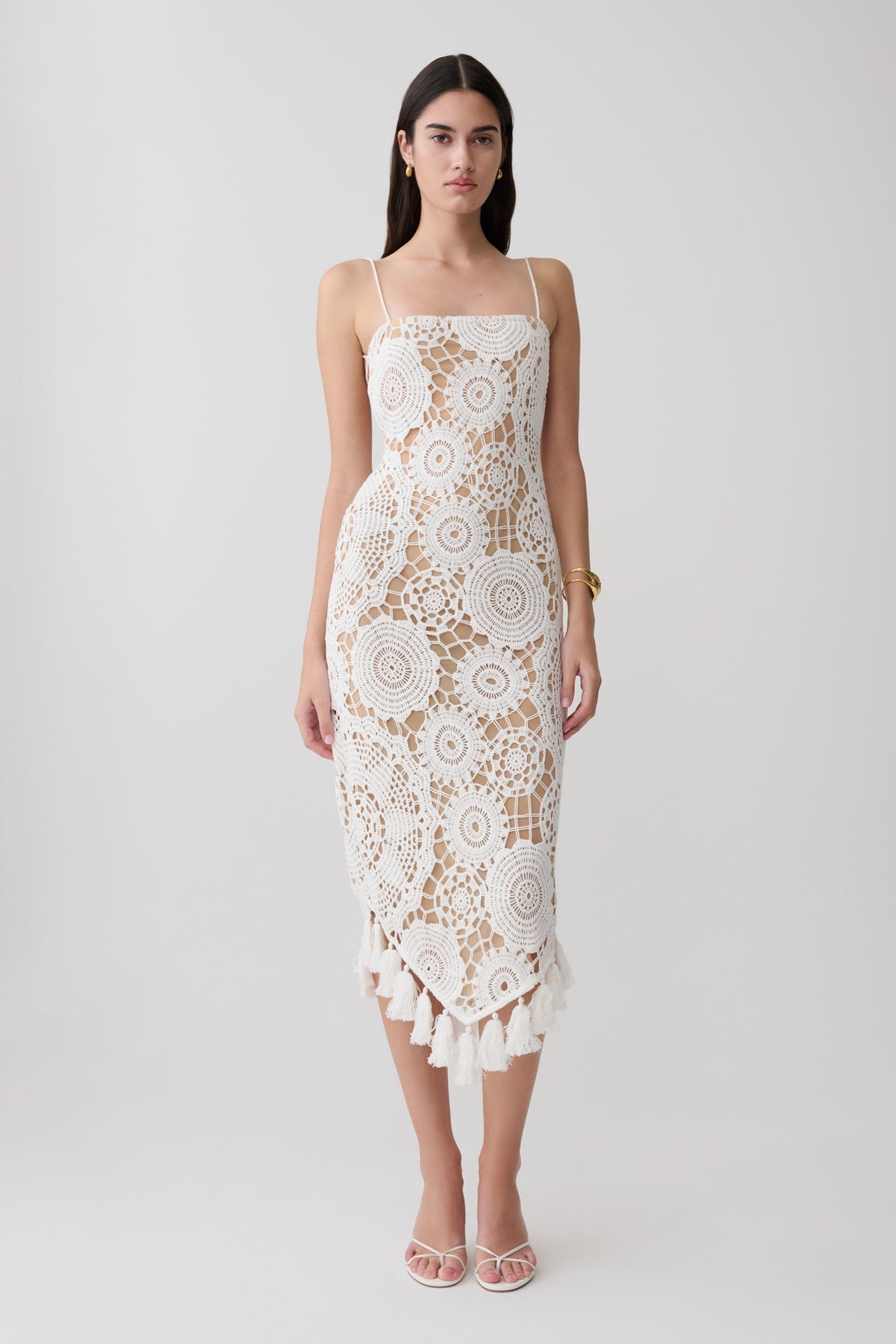 A woman modeling a white crochet lace midi dress with spaghetti straps, a fitted silhouette, and a ruffled hem, holding a small beige clutch against a light gray studio background.