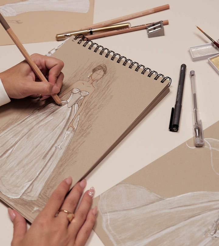 Person drawing a figure in a sketchbook with art supplies on a table.