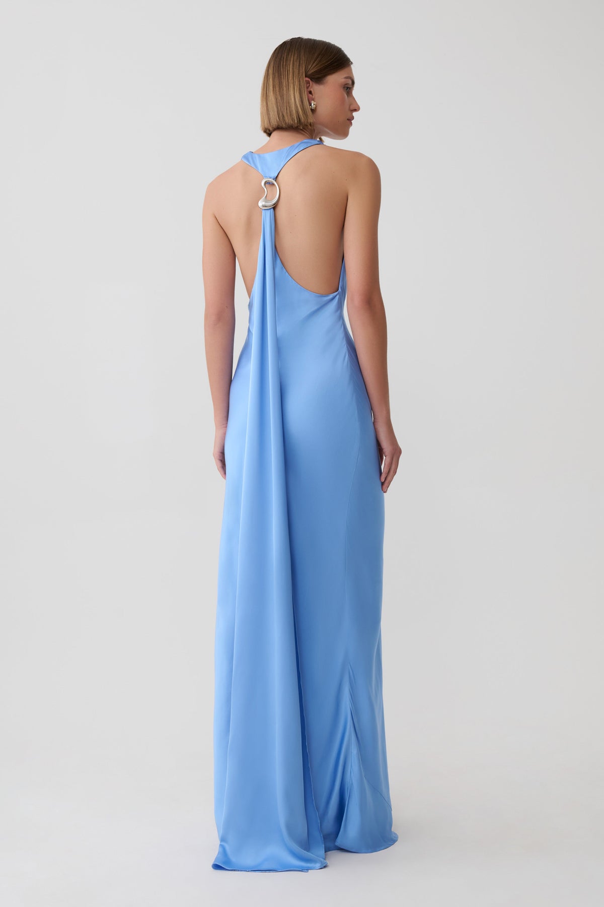A woman shown from the back wearing a light blue satin maxi dress with a halter-style open back and a prominent gold O-ring detail at the neckline, set against a plain light gray studio background.