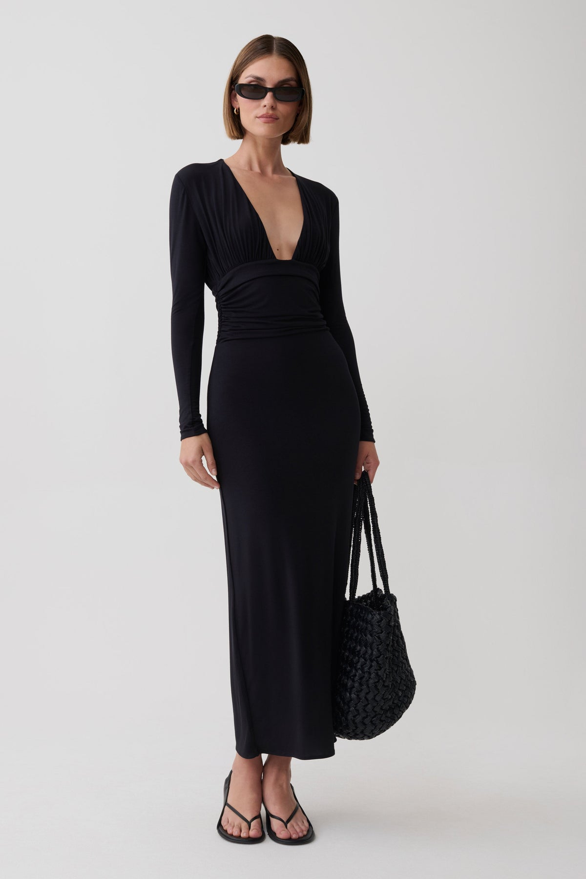 A woman modeling a long-sleeve black maxi dress with a deep V-neck and gathered waist, styled with black sunglasses, black strappy sandals, and a black woven handbag against a light gray studio background.