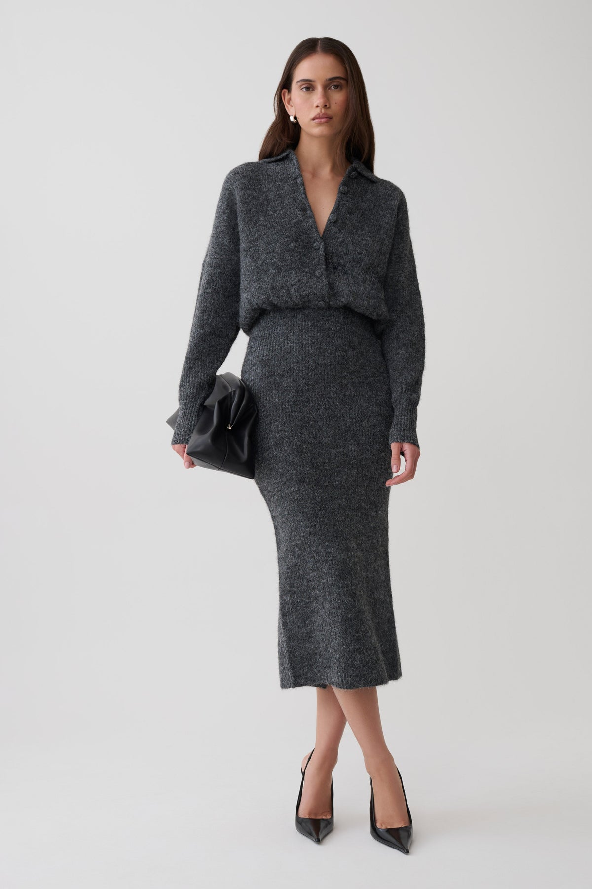 A woman modeling a dark gray knit long-sleeve dress, styled with black pointed-toe heels and a black clutch bag against a light gray studio background.