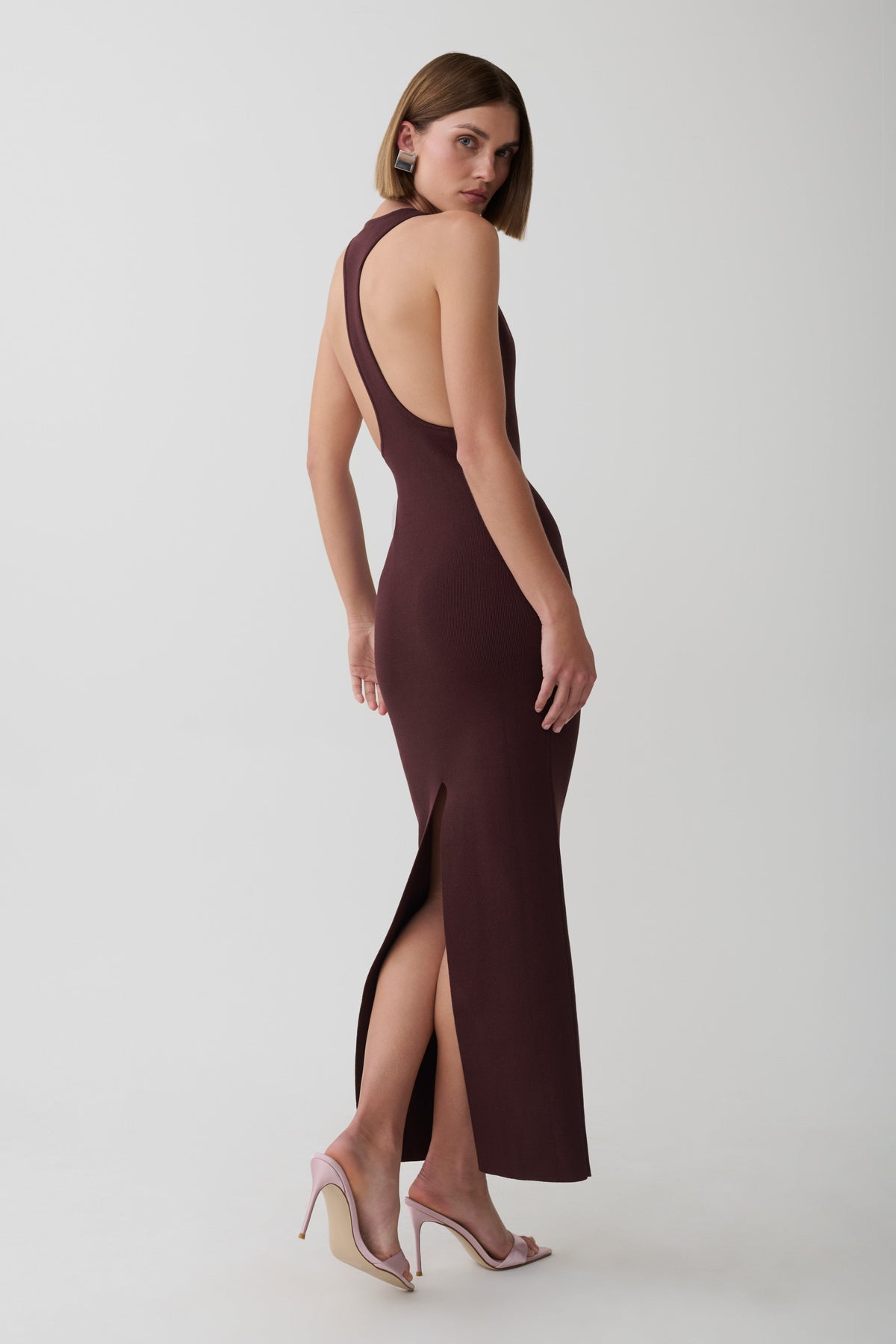 A woman modeling a sleeveless, backless, floor-length dark plum maxi dress with a high side slit, paired with clear high-heel sandals, against a light gray studio background.