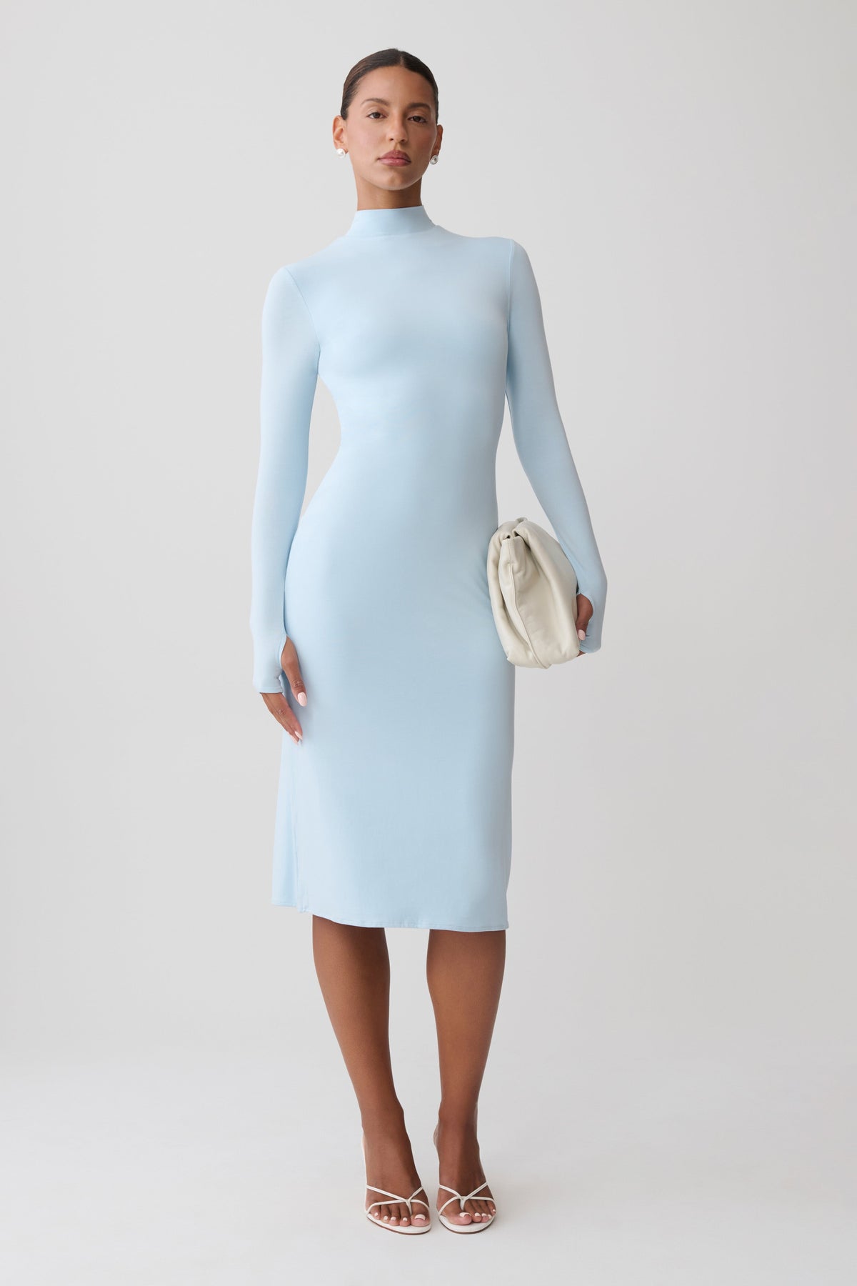 A model stands against a light gray studio background wearing a fitted light blue mock-neck midi dress with long sleeves, holding a small cream clutch and wearing clear-strap high-heel sandals.
