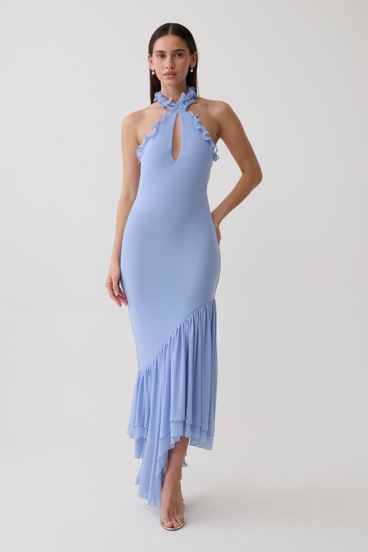 A woman modeling a light blue halter-neck maxi dress with ruffle trim and an asymmetrical ruffled hem, photographed against a plain light gray studio background.