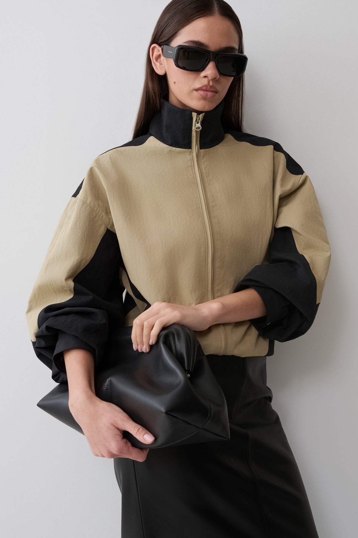 A model wearing a beige and black color-blocked zip-up jacket, black sunglasses, and a black skirt, holding a black leather clutch against a light gray background.