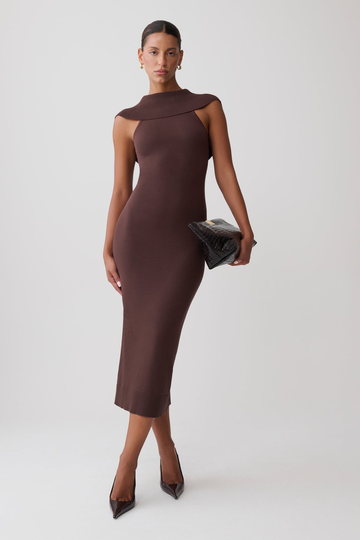 A model wearing a sleeveless, bodycon midi dress in a dark chocolate brown color, styled with a black textured clutch and dark pointed-toe heels, set against a plain light gray studio background.