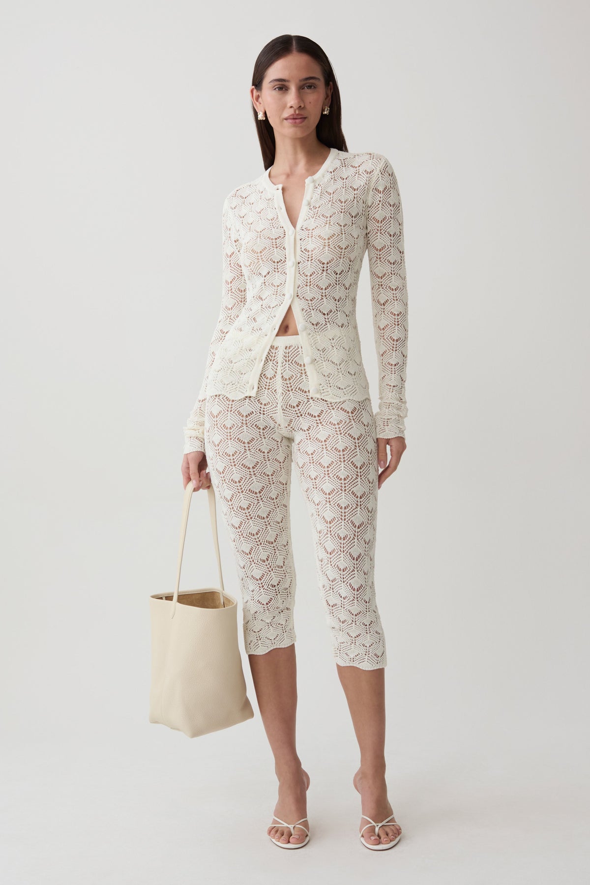 A model stands against a light gray studio background wearing a matching cream lace three-piece set consisting of a long-sleeve button-front cardigan, cropped capri pants, and a tie belt. She is holding a cream canvas tote bag and wearing strappy high-heeled sandals.