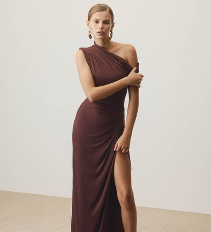 Woman wearing a burgundy one-shoulder gown with a slit on a plain background.