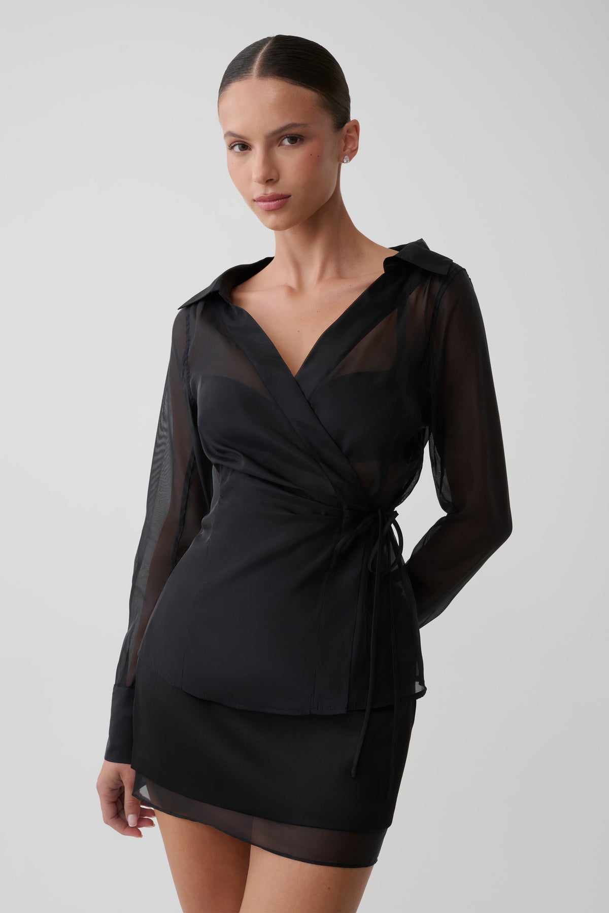 A model wearing a black long-sleeve wrap dress with sheer mesh panels and a self-tie waist, photographed against a neutral light gray studio background.