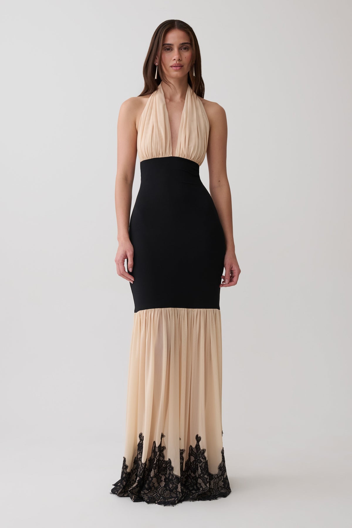 A woman modeling a sleeveless halter-neck maxi dress with a black fitted waist and skirt, and a sheer beige lower skirt with black lace trim at the hem, against a light gray studio background.