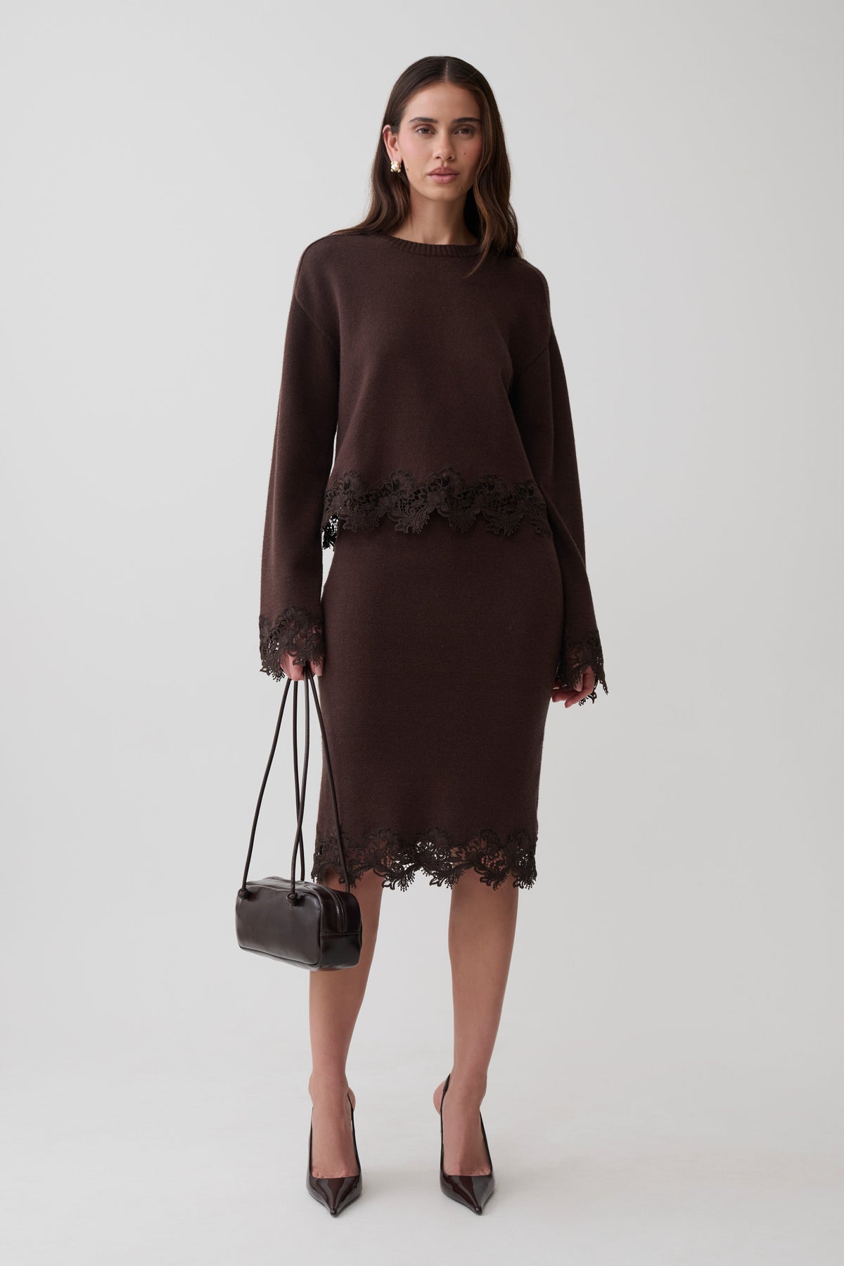 A woman models a dark brown long-sleeve knit jumper and midi skirt with dark brown lace trim, holding a small black handbag against a light gray studio background.
