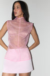 A woman modeling a sleeveless pink sheer mesh top with a high mock neck and a light pink mini skirt, set against a plain light gray studio background.