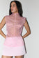 A woman modeling a sleeveless pink sheer mesh top with a high mock neck and a light pink mini skirt, set against a plain light gray studio background.