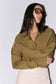 A woman wearing a relaxed-fit olive green knit wrap sweater and cream-colored bottoms, styled with oversized black sunglasses, posed against a plain light gray background.