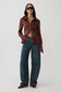 A model stands against a light gray studio background wearing a brown long-sleeve button-up shirt, blue wide-leg jeans, dark sunglasses, and dark pointed shoes.