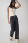 A woman modeling a pair of dark gray wide-leg jeans with contrast light stitching, styled with a cream ribbed tank top, a navy baseball cap, and white heels against a plain light gray studio background.