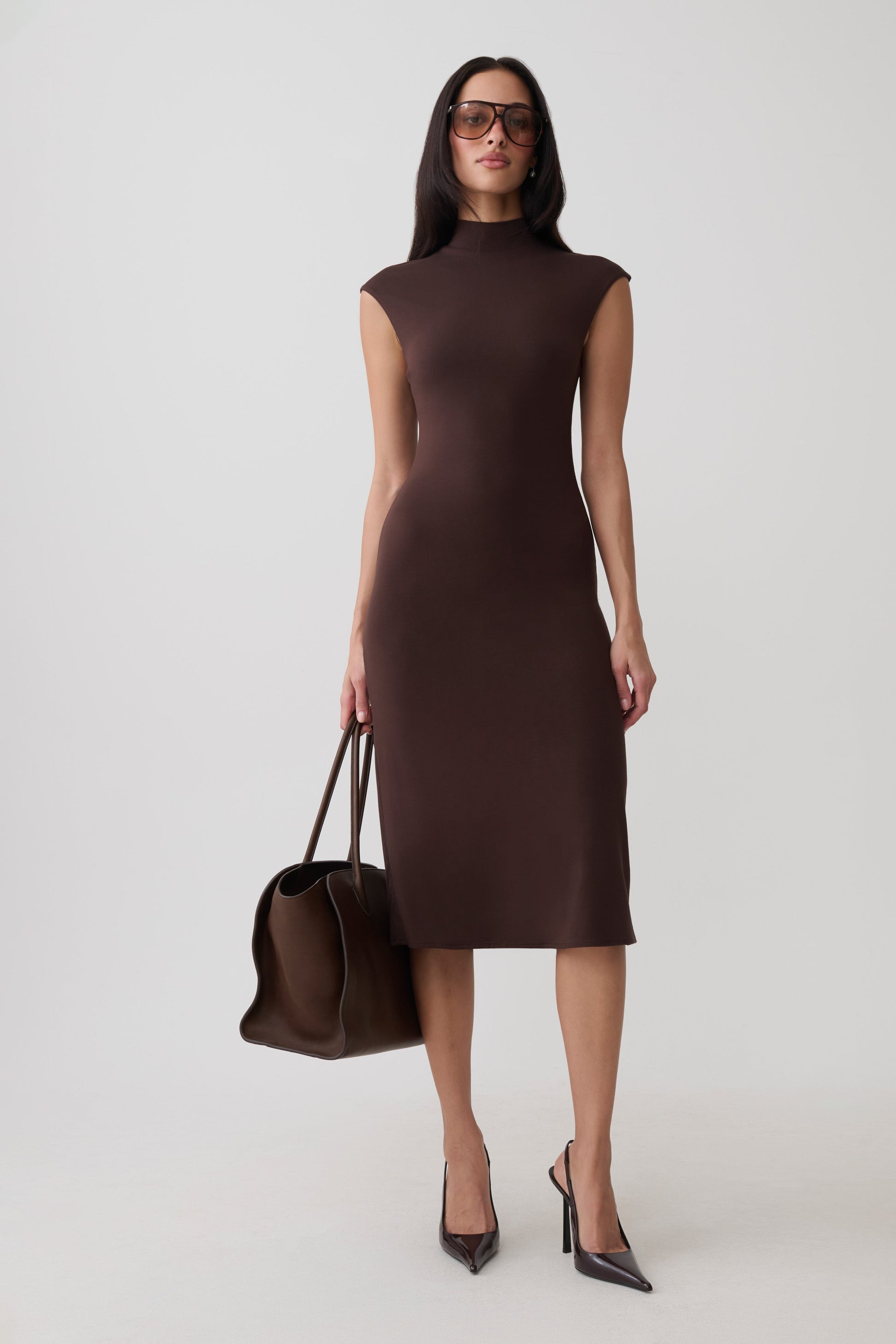 A woman modeling a sleeveless, high-neck, knee-length brown dress, holding a matching brown tote bag and wearing dark pointed-toe heels against a light gray studio background.