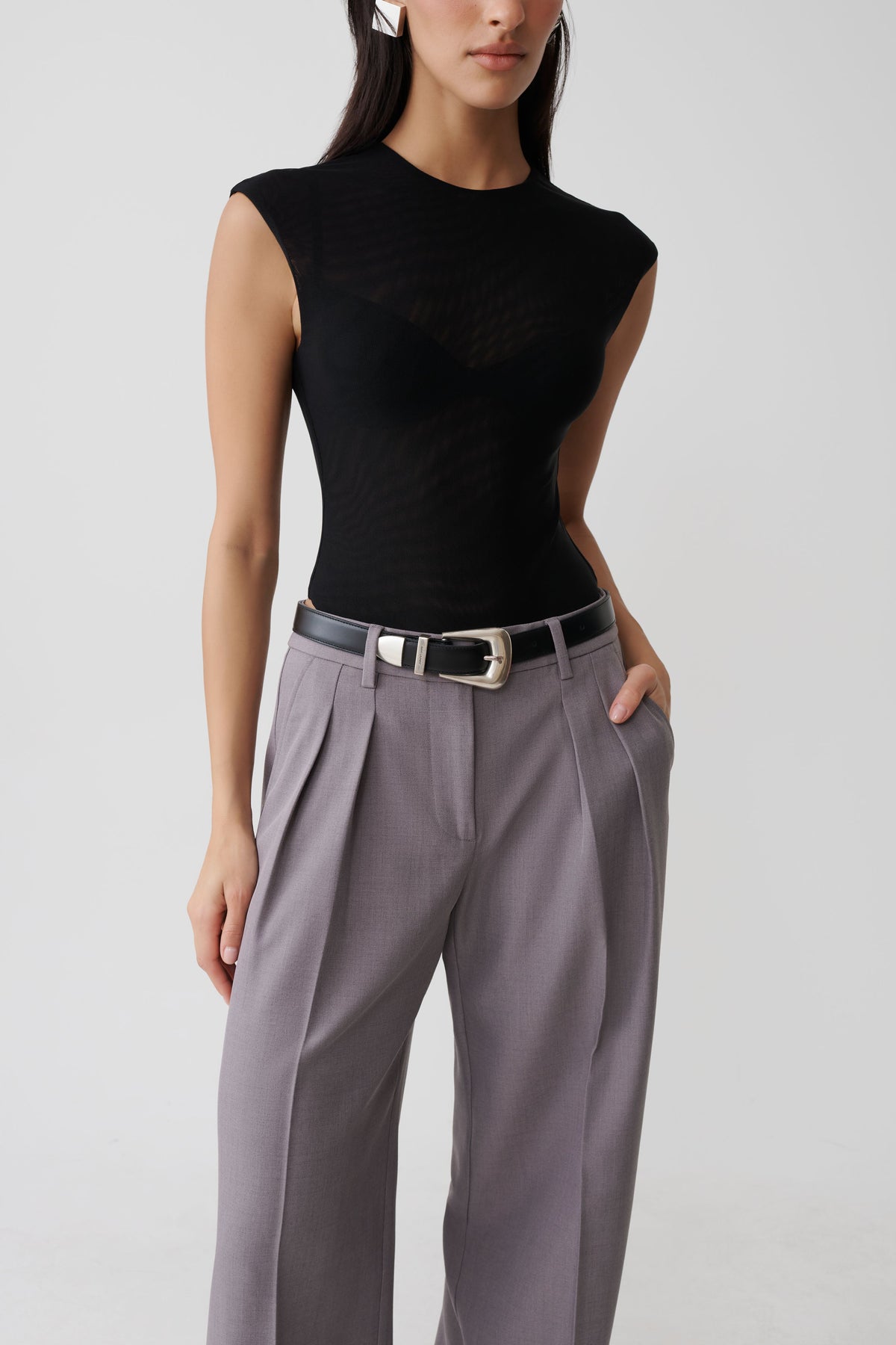 A model wearing a black sleeveless textured top and high-waisted gray pleated trousers with a black belt featuring a silver buckle, shot against a light gray studio background.
