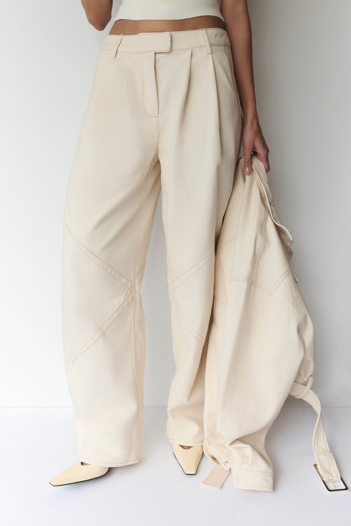 A model wearing high-waisted, wide-leg cream trousers with diagonal seam details, holding an extra pair of the same trousers against a plain white studio background.