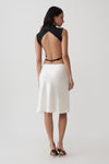 A woman shown from the back wearing a sleeveless black top with an open back and a white knee-length skirt, photographed against a plain light gray studio background.
