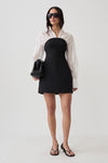 A model stands against a light gray studio background wearing a black strapless mini dress with a white long-sleeve collared shirt layered over it, black sunglasses, black sandals, and holding a black handbag.
