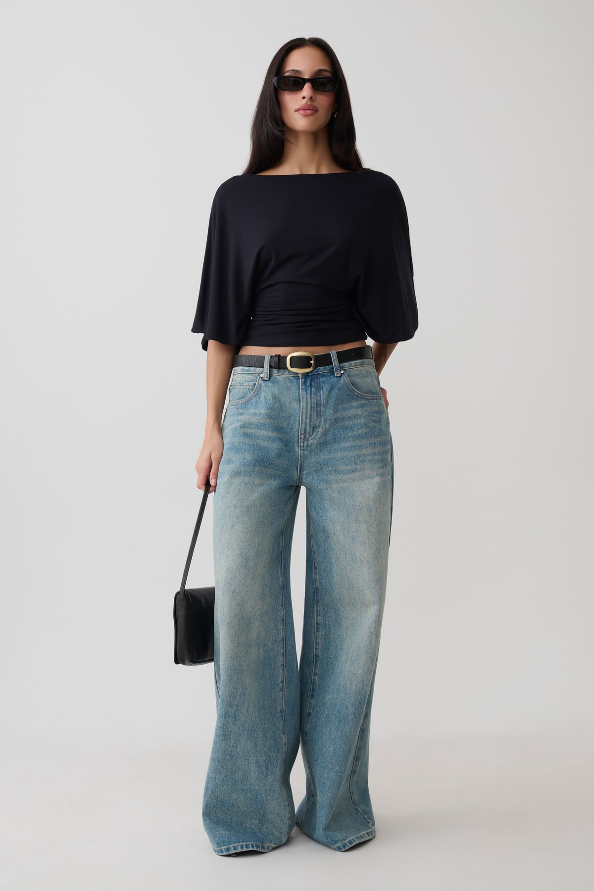 A woman modeling a black boat-neck top with elbow-length sleeves and light-wash wide-leg jeans, styled with a black belt, sunglasses, and a small black handbag against a light gray studio background.