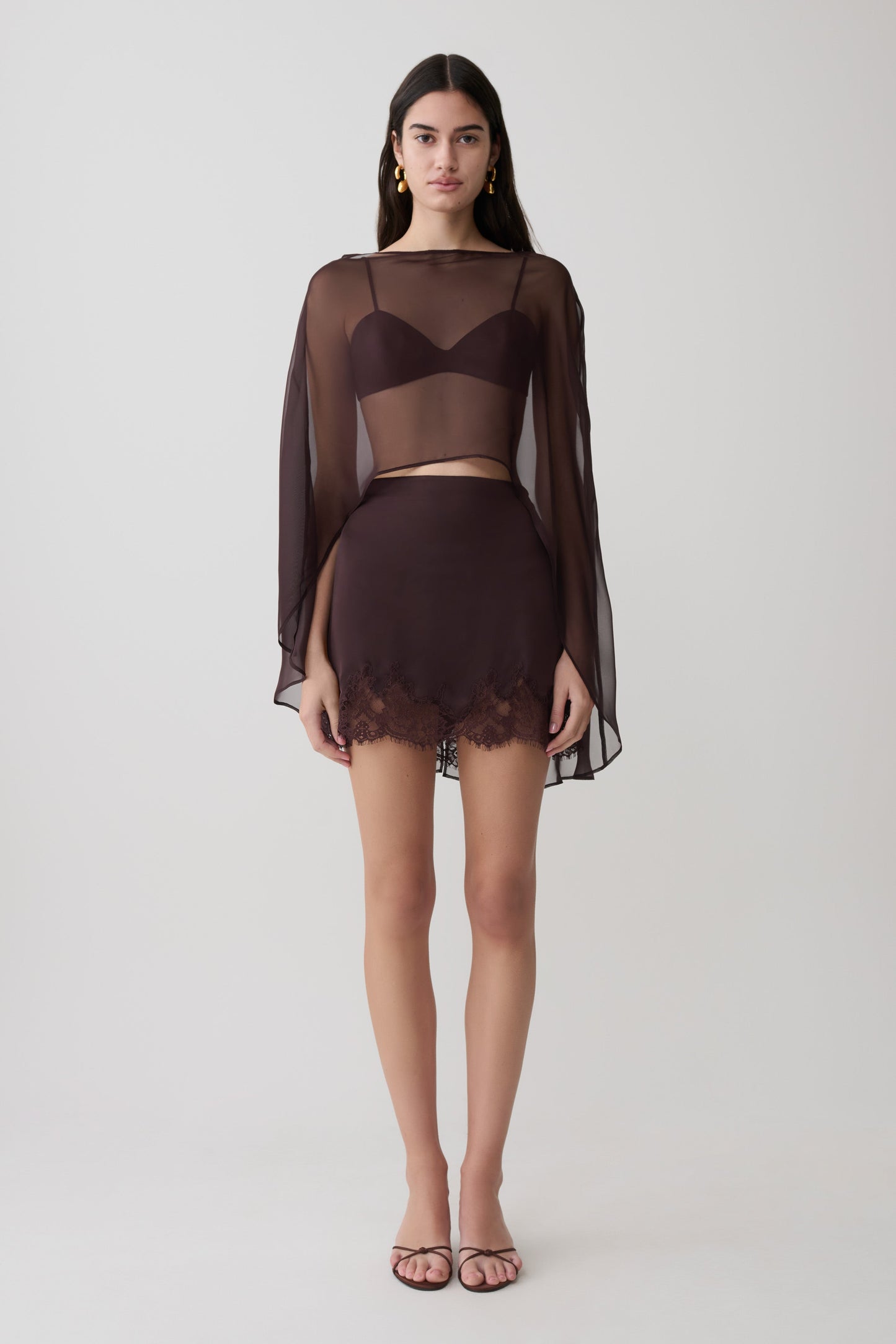 Woman wearing brown bralette with chiffon cape with matching lace and satin mini skirt.