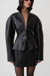 A model wearing a black textured faux-leather jacket with a high collar and a matching black mini skirt, photographed against a light gray studio background.