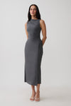 A woman modeling a sleeveless, dark gray midi dress with a draped neckline and side tie detail, paired with white strappy heels, against a light gray studio background.
