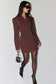 A woman modeling a dark brown blazer dress with structured shoulders, paired with black tights, pointed-toe heels, and a small matching handbag against a plain light gray background.