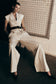 A model wearing natural beige mid-rise wide leg linen pants with a belt, sitting against a tiled wall.