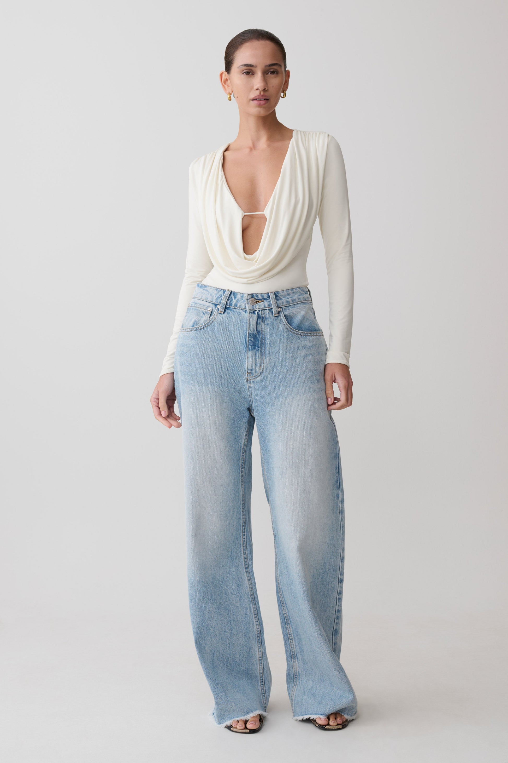 Woman wearing a white long-sleeve top and light blue jeans on a plain background