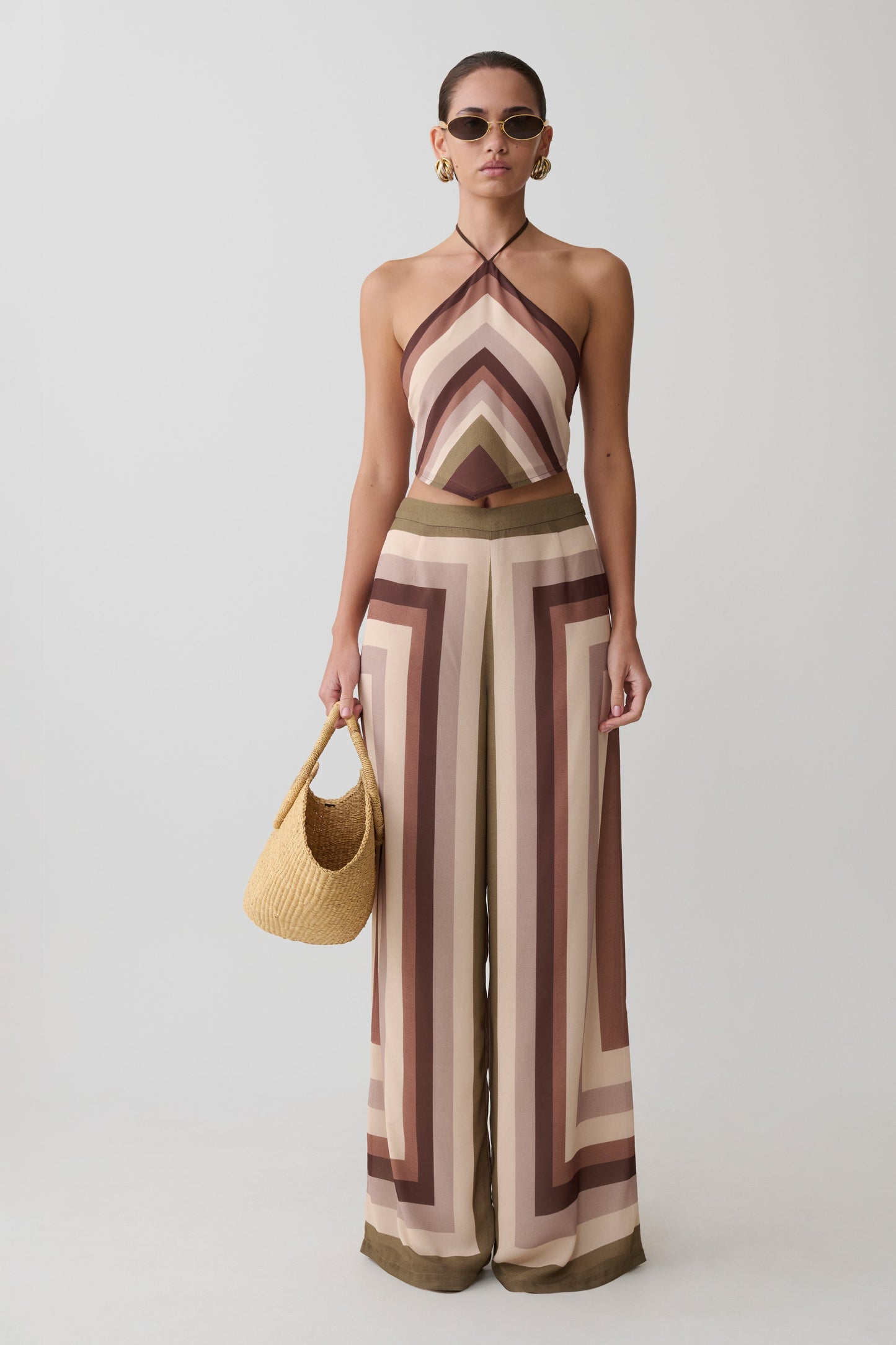 Woman wearing a patterned jumpsuit holding a straw bag on a plain background