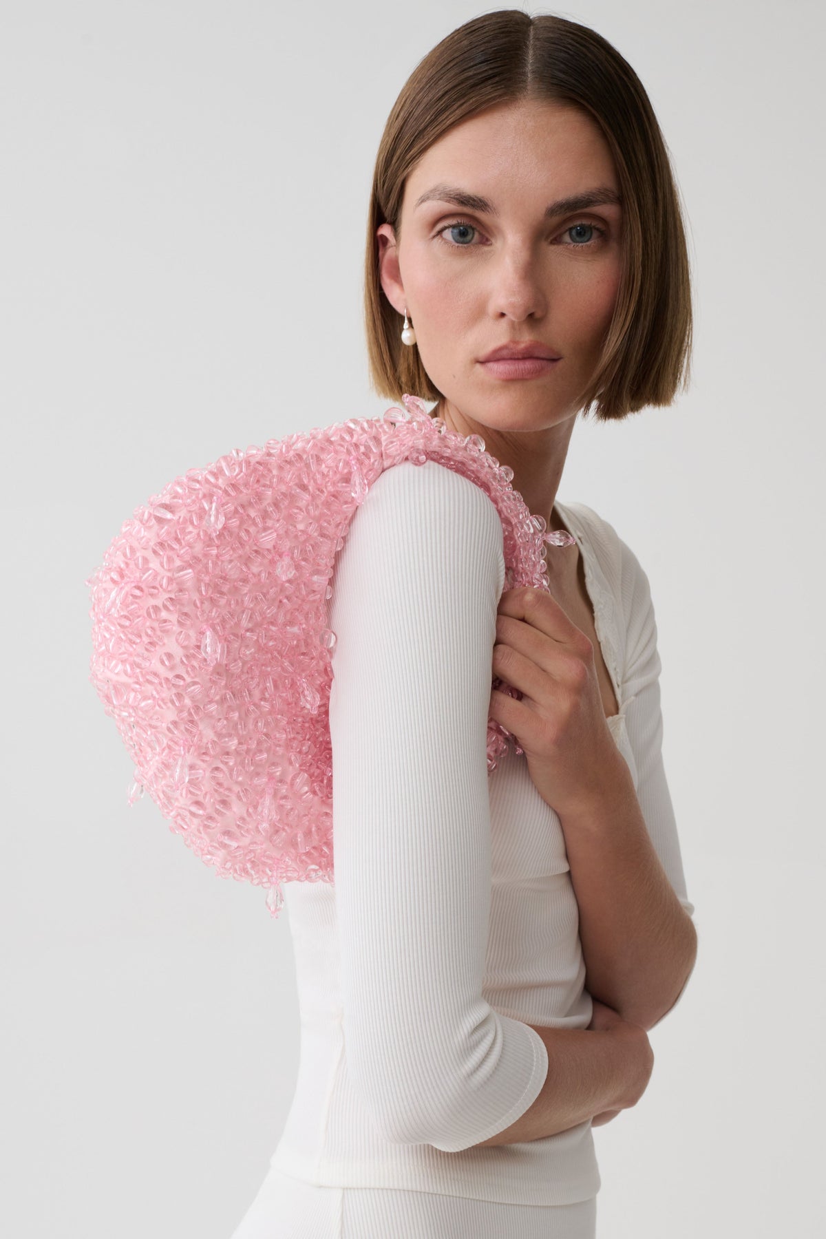 A model wearing a white ribbed long-sleeve top holds a large, round, light pink beaded bag against her shoulder, photographed against a clean light gray studio background.