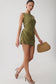 A woman models a sleeveless olive green mini dress with gathered draping at the skirt, paired with brown heeled sandals and a woven straw handbag, against a plain light gray studio background.