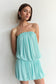 A woman wearing a strapless light aqua blue mini dress with a gathered, tiered ruffled design, posing against a plain off-white wall.