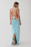 A woman modeling a light blue maxi dress from the back, showing an open back with thin straps and a draped skirt, against a plain light gray studio background.