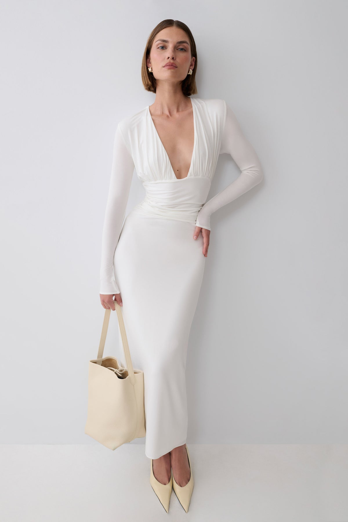 A woman models a fitted white long-sleeve midi dress with a deep plunging neckline and a ruched waist, paired with cream pointed-toe heels and a cream tote bag against a light gray studio background.