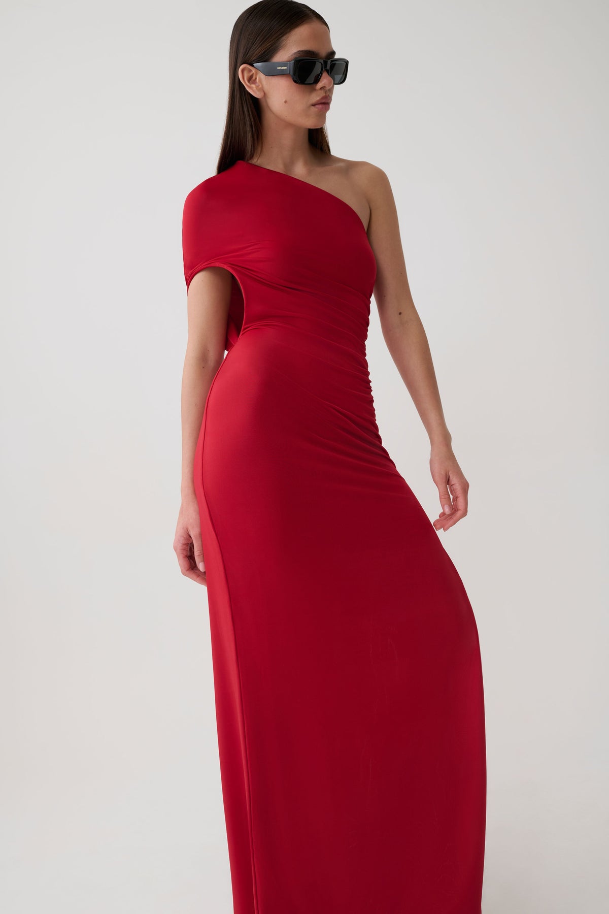 A woman modeling a floor-length, one-shoulder red dress with a side cutout, photographed against a plain light gray studio background.