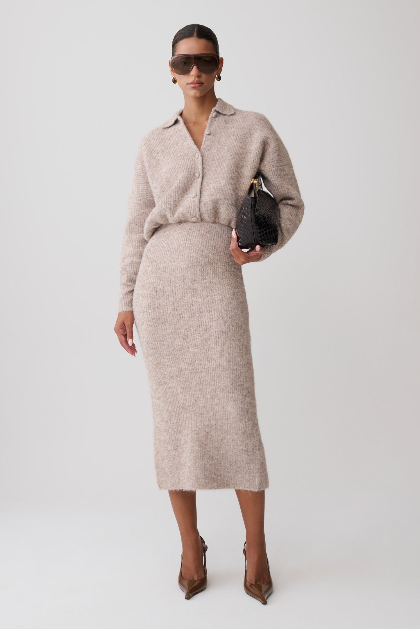 A model wearing a light beige knit long-sleeve collared midi dress, styled with dark sunglasses, a small black handbag, and brown pointed-toe heels against a light gray studio background.