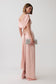 A woman modeling a floor-length blush pink gown with draped one-shoulder styling, an open back, and a side slit, holding a silver clutch against a light gray studio background.