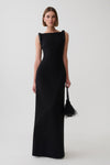 A model wearing a sleeveless black maxi dress with structured shoulders and holding a black feather-trimmed handbag against a light gray studio background.