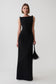 A model wearing a sleeveless black maxi dress with structured shoulders and holding a black feather-trimmed handbag against a light gray studio background.