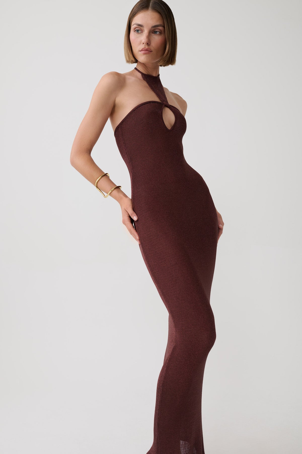A woman modeling a fitted halter-neck bodycon dress in a deep burgundy or maroon knit, featuring a keyhole cutout at the bust. She is posed against a clean, light gray studio background.