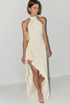 A model wearing a cream-colored halter-neck maxi dress with a high-low ruffled hem and a matching draped train, styled with gold accessories against a plain light gray studio background.